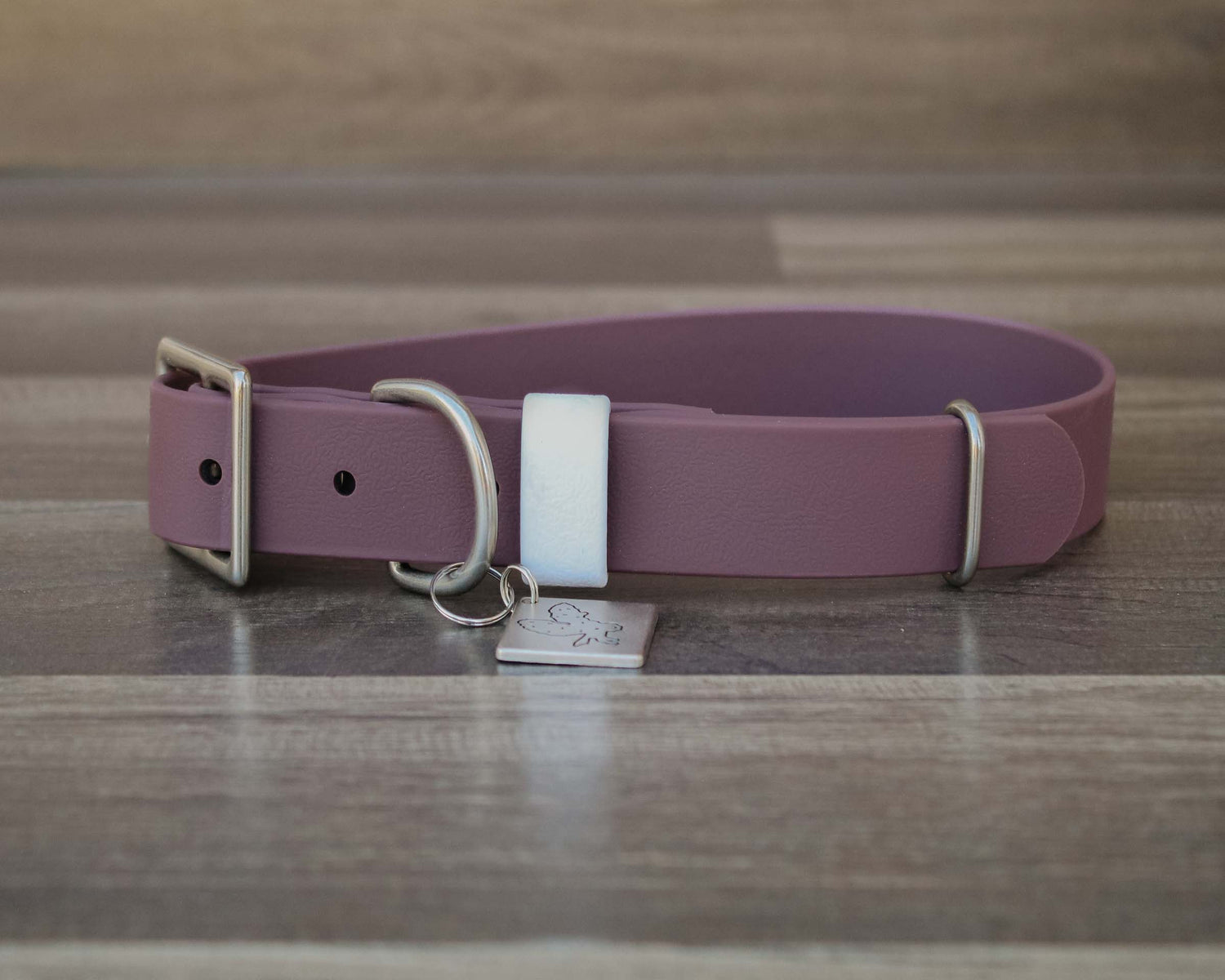 A dusty purple leather dog collar with silver hardware, a white plastic keeper, and a small engraved metal ID tag, displayed on a wooden surface.