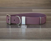 A dusty purple leather dog collar with silver hardware, a white plastic keeper, and a small engraved metal ID tag, displayed on a wooden surface.